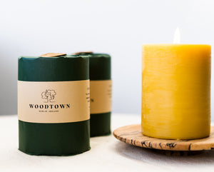 A pillar candle of pure, yellow coloured, Irish beeswax sitting on a spalted beech wooden candle plate. Beside sit 2 wrapped pillar candles, one in front of the other, in dark green paper and a cream coloured belly band with the Woodtown logo on