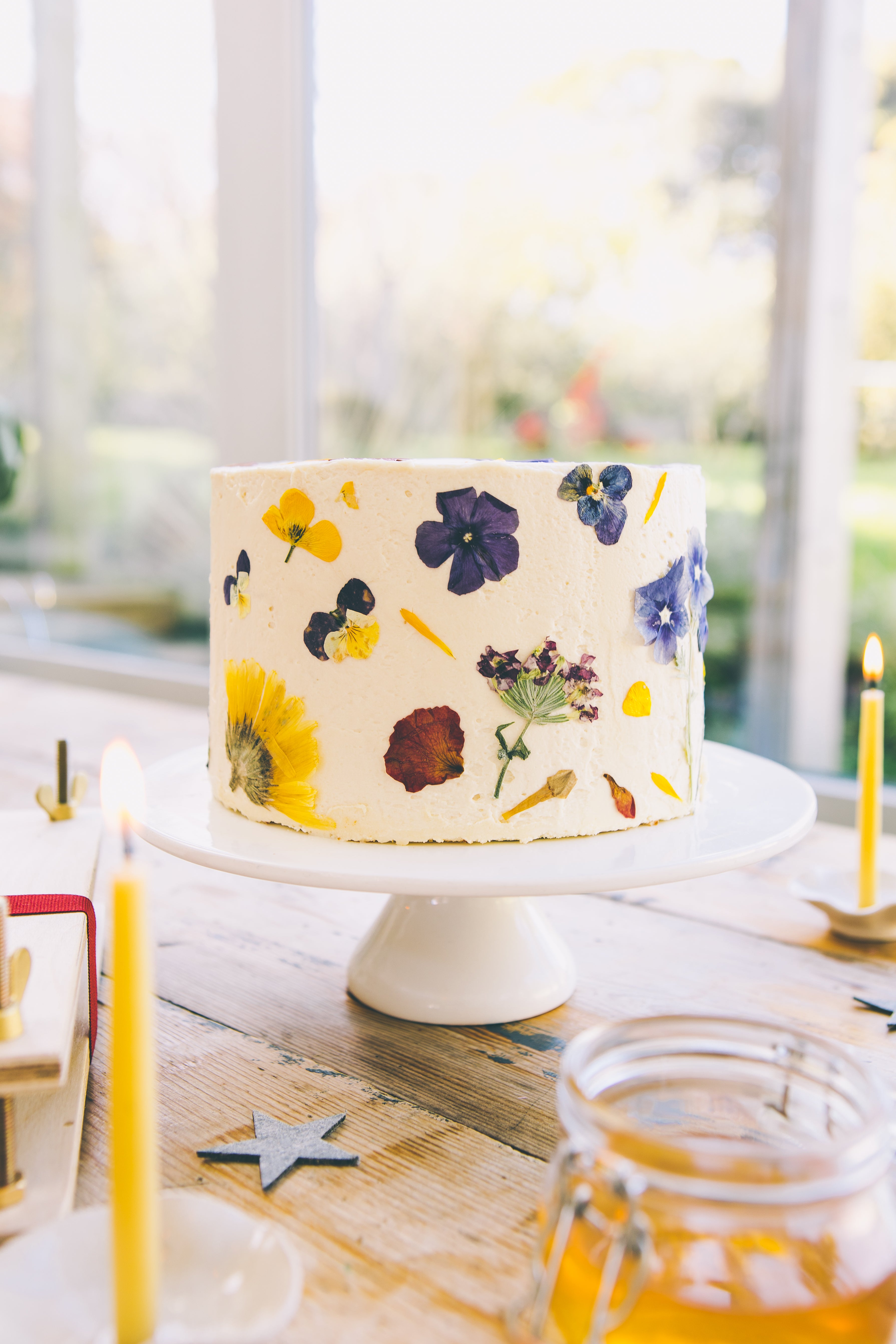 A large white buttercream frosted cake decorated in pressed wild flower in various colours, with lit, yello beeswa candles in the foreground and background.