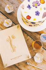 Load image into Gallery viewer, A white buttercream frosted cake decorated with various pressed wild flowers in different colours. Cake is sitting on a wooden table, beside a plywood flower press, engraved with Irish wild flowers. A jor of open honey and honey dripper sit on the table too. A few lit beeswax candles sit lit in amongst the press and cake.
