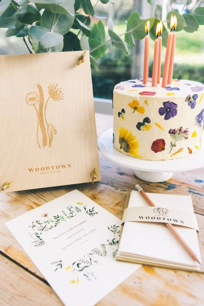 A display on a wooden table of a white swiss meringue buttercream frosted cake decorated with various coloured pressed wildflower, beside a plywood flower press standing upright beside it. Some cream paper notelets and pencil sit in the foreground. Woodtown Studio and Camerino bakery collaboration