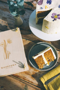 A photo taken looking down upon a woodtown studio birch plywood flower press, besid a slice of cake, and a buttercream frosted cake decorated in pressed wild flowers in various colours.