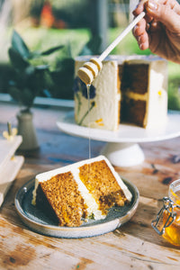 A hand holding a honey dripper drizzling honey over a cut slice of songe cake, with a white buttercream frosted cake decorated with pressed wild flower in the background
