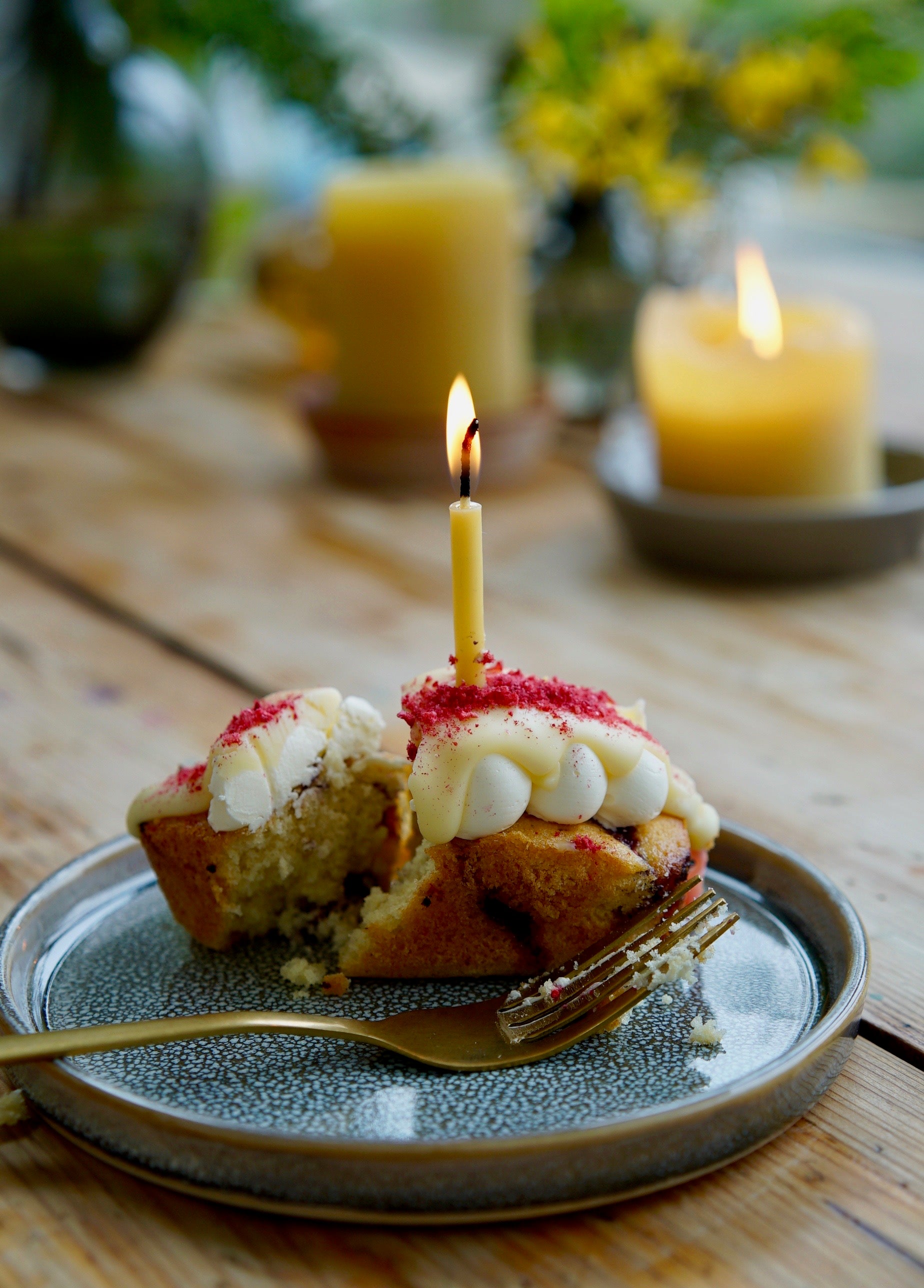 An image of a woodtown studio bee happy beeswax candle lit atop an iced cake on a plate with a bronze fork, all sitting on a wooden table