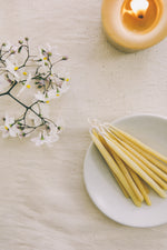 Load image into Gallery viewer, a white dish with a dozen yellow beeswax celebration candles on. A lit yellow beeswax pillar candle lit, half out of shot. Some yellow daisies, all of these lying on a cream cotton fabric
