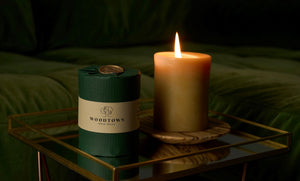 A wrapped woodtown studio beeswax pillar candle, in dark green wrap and a cream belly band with text,  beside an unwrapped and burning beeswax pillar candle. Both beeswax pillar candles are on a mirrored tray, infront of a dark green sofa. The lit candle is sitting on a spalted beech flat plate