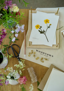 A top down view of the Woodtown forager's flower press on a table surrounded by twine, scissors, pencil, cards, flowers in bud vases, cotton bag with Woodtown branding on it. A pressed yellow buttercup is laying on the flower press
