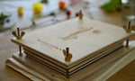 Load image into Gallery viewer, Birch plywood flower press laying on a wooden table. Woodtown logo engraved on the top board. Engraving of flowers on top board are out of focus. Brass wingnuts are in focus
