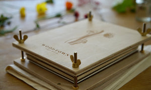 Birch plywood flower press laying on a wooden table. Woodtown logo engraved on the top board. Engraving of flowers on top board are out of focus. Brass wingnuts are in focus