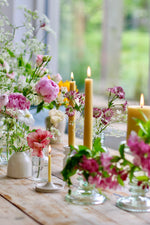 Load image into Gallery viewer, A few yellow beeswax candles lit, in candleholders, on a wooden table with pink and white blooms scattered in various size vases amongst them. Some glass holders and white ceramic holders are used. Pink peopnies, and cow parsley flowers
