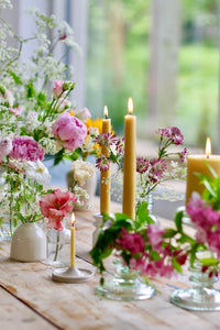 A few yellow beeswax candles lit, in candleholders, on a wooden table with pink and white blooms scattered in various size vases amongst them. Some glass holders and white ceramic holders are used. Pink peopnies, and cow parsley flowers
