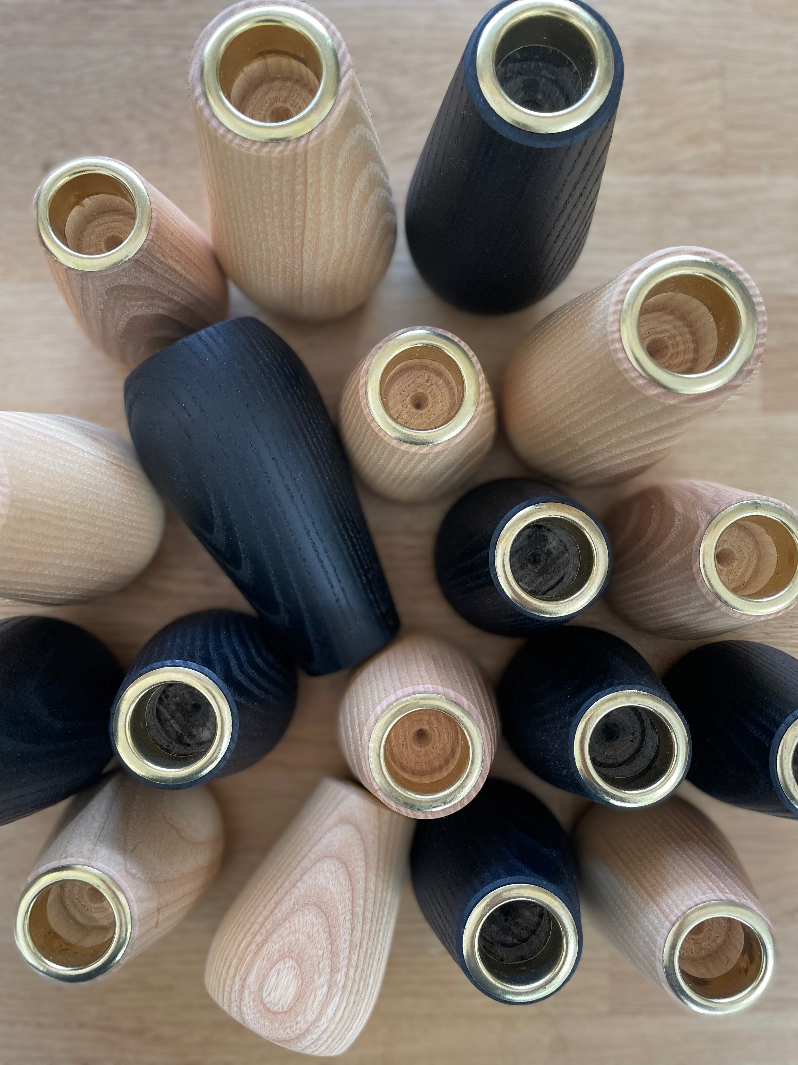 18 hand-tuened wooden candlestick holders grouped together on a table, with one lying on its side. A mix of natural ash wood and black ebonised ash. Each has a brass component insert where the candle sticks into.