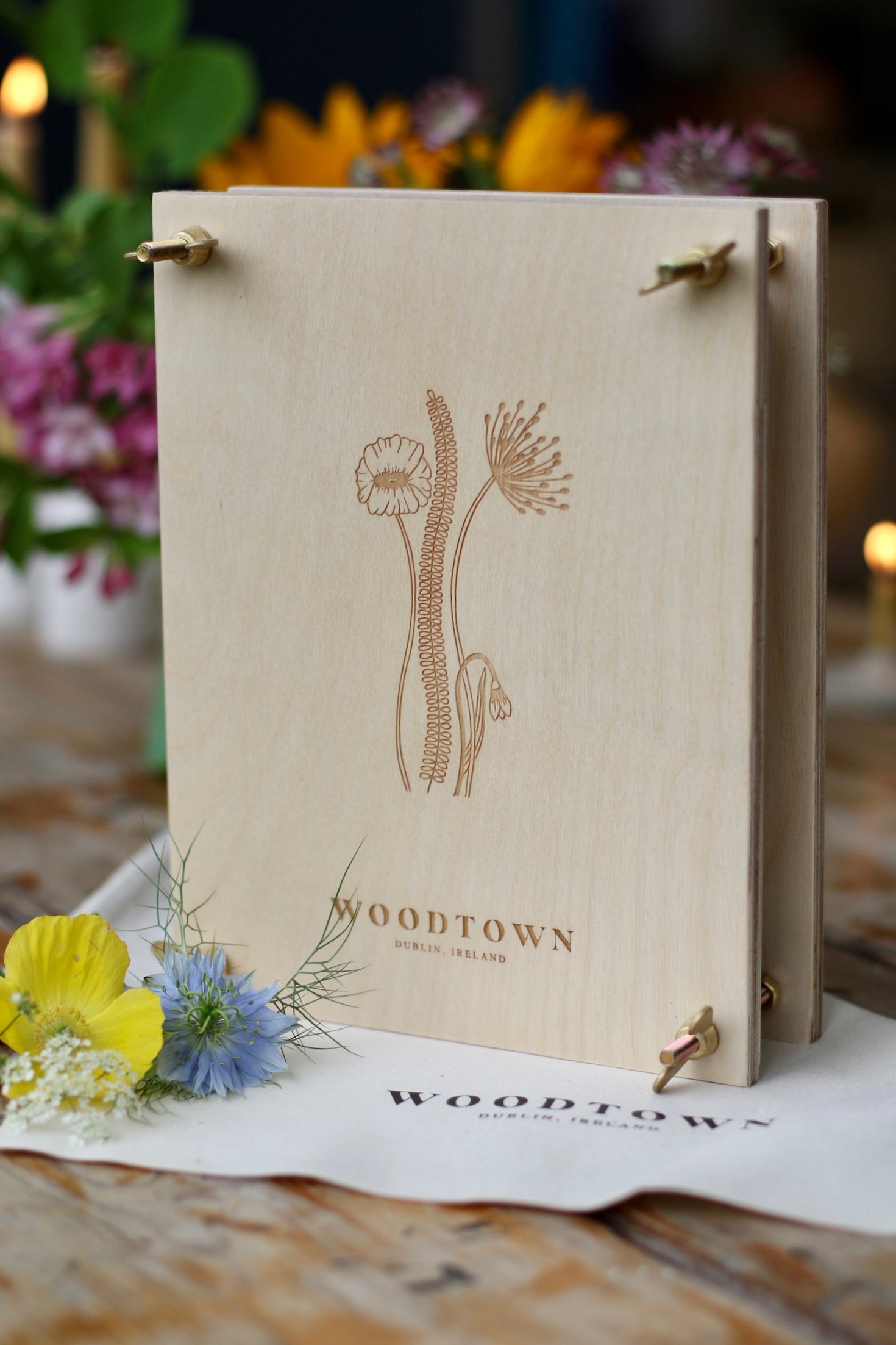 A birch plywood flower press, standing upright, with  3 tall Irish wildflower and 1 small flower engraved on the front. Brass hardware are used to bolt it together. Brass wingnuts. Background are multi colour flowers