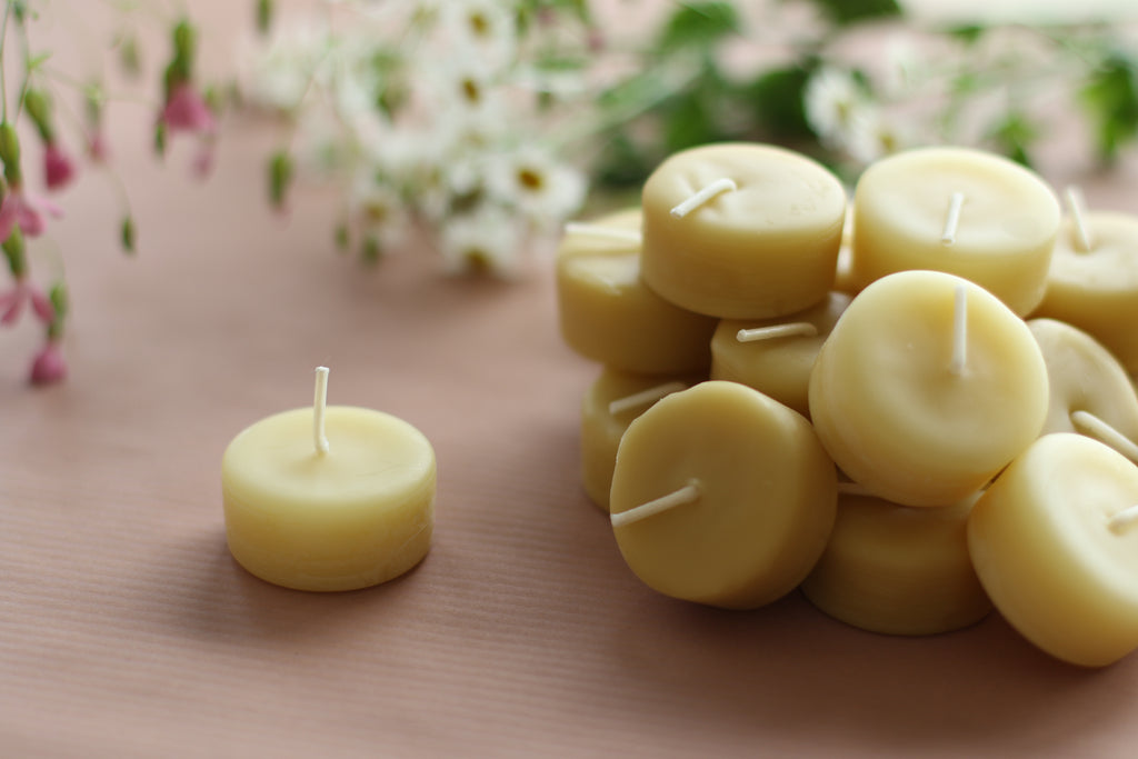 a pile of 18 pure beeswax tea lights with one placed out on its own, sitting on a pale blush pink table top and flowers in the background