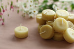 Load image into Gallery viewer, a pile of 18 pure beeswax tea lights with one placed out on its own, sitting on a pale blush pink table top and flowers in the background
